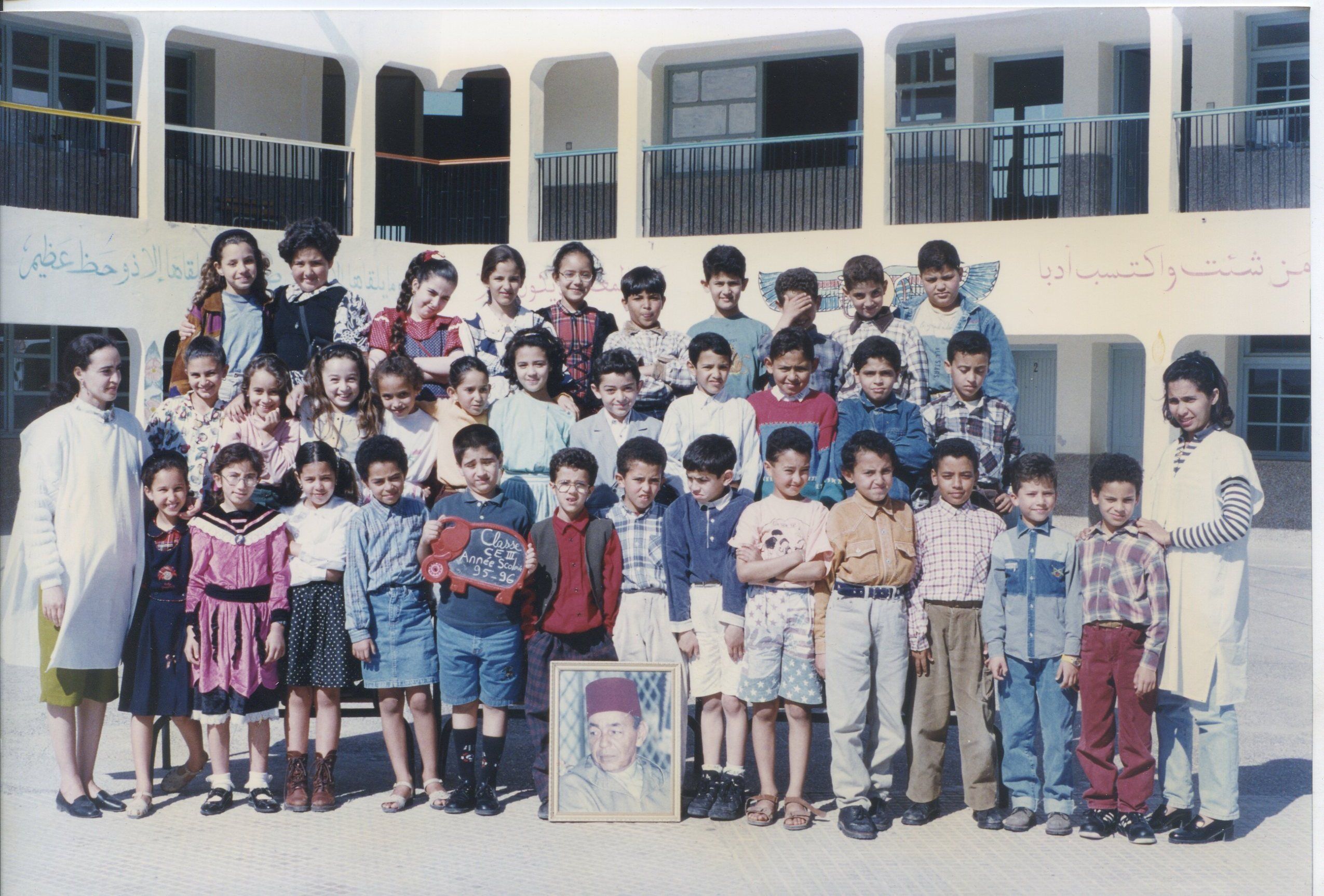 Photo de classe en couleur, tirage argentique : une quarantaine d'enfants et leurs enseignantes posent dans la cour, devant les arcades blanches de l'école.