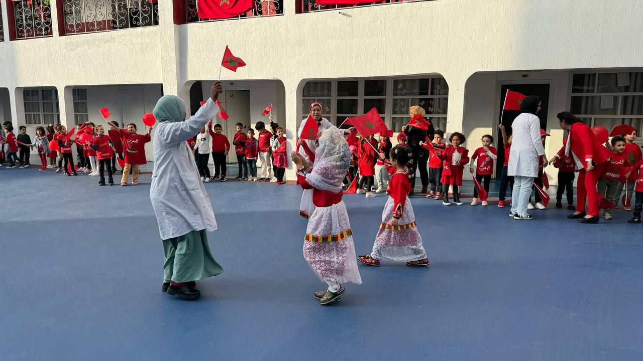 Dans la cour, de jeunes élèves vêtus de rouge agitent des drapeaux marocains ; deux fillettes en tenue traditionnelle mènent la ronde.